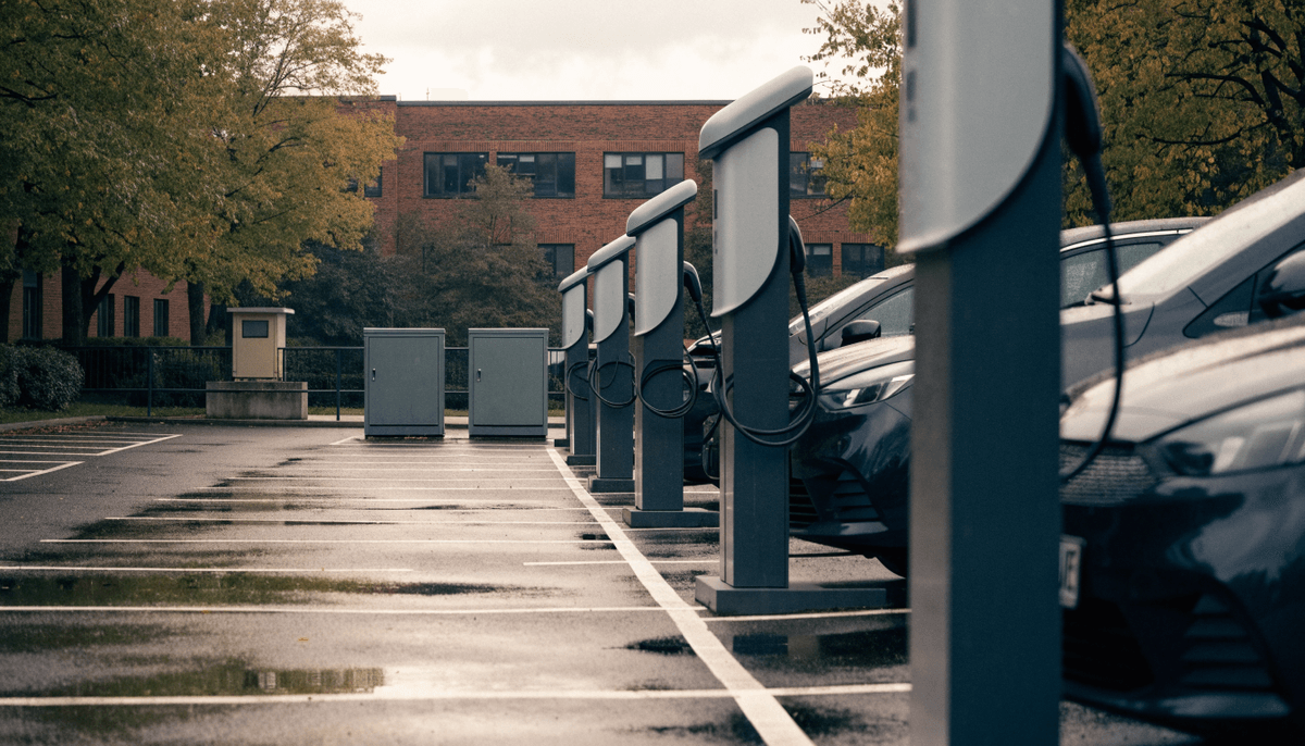 A UK business car park on an overcast autumn afternoon with a row of pedestal EV chargers, compact grey-green equipment cabinets and a small transformer kiosk on a concrete plinth behind them, wet tarmac and a red-brick building beyond
